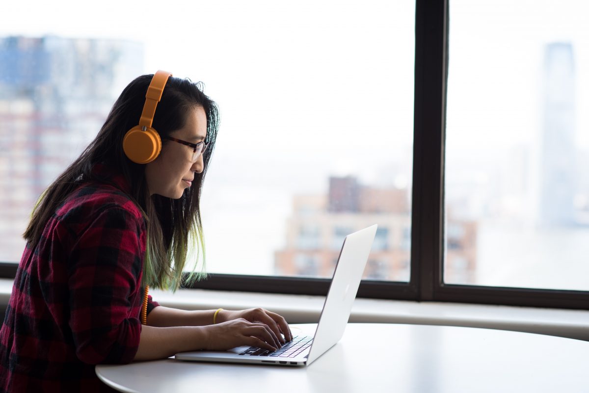 Asian American woman looks at her laptop while wearing headphones. She is seated in front of a window and a cityscape.