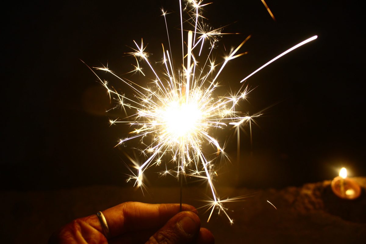 A hand holds a sparkler that is sending out a halo of sparks in front of a dark background.