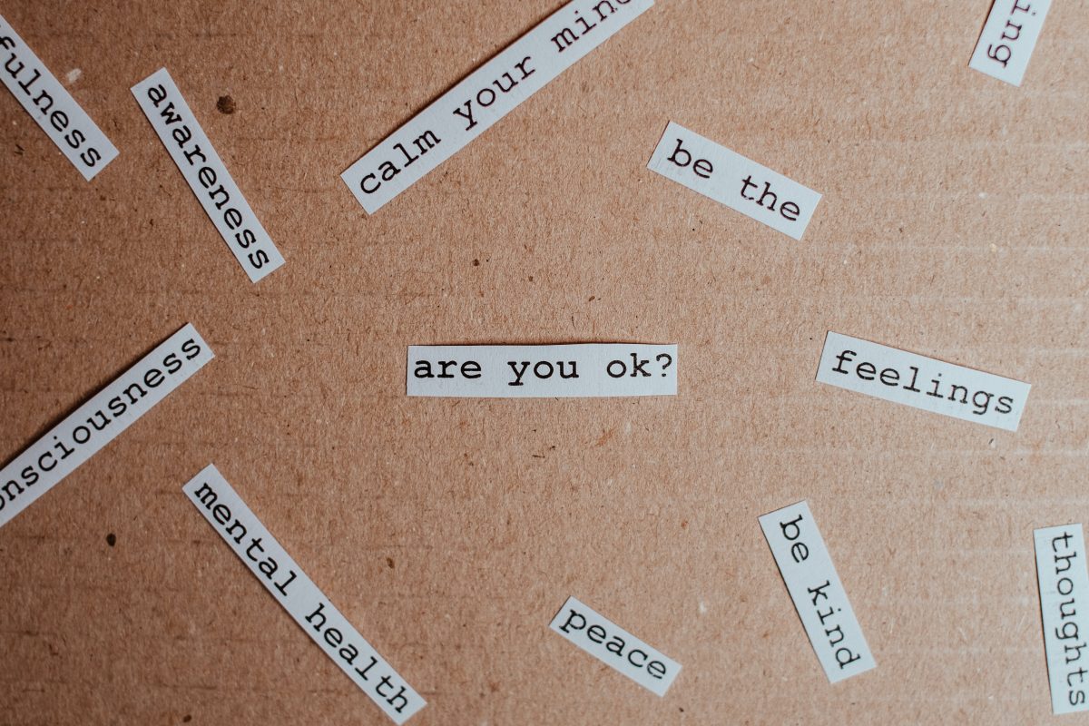 Small strips of white paper with phrases like "are you ok," "be kind," and "awareness" are scattered across a brown cardboard background.