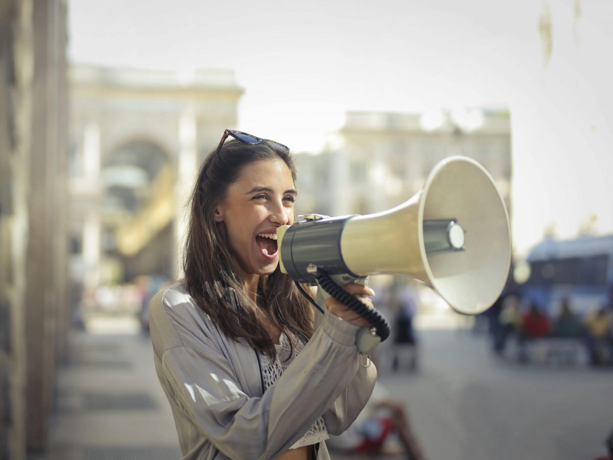 A young woman with straight brown hair and tan skin stands in a city street, holding a megaphone as she speaks.