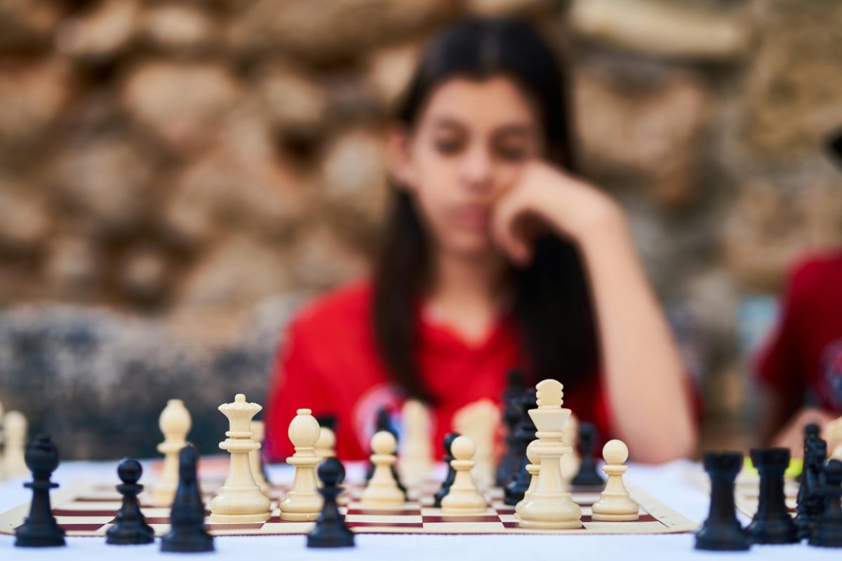 A young woman with long brown hair is blurred out in the background behind a game of chess.