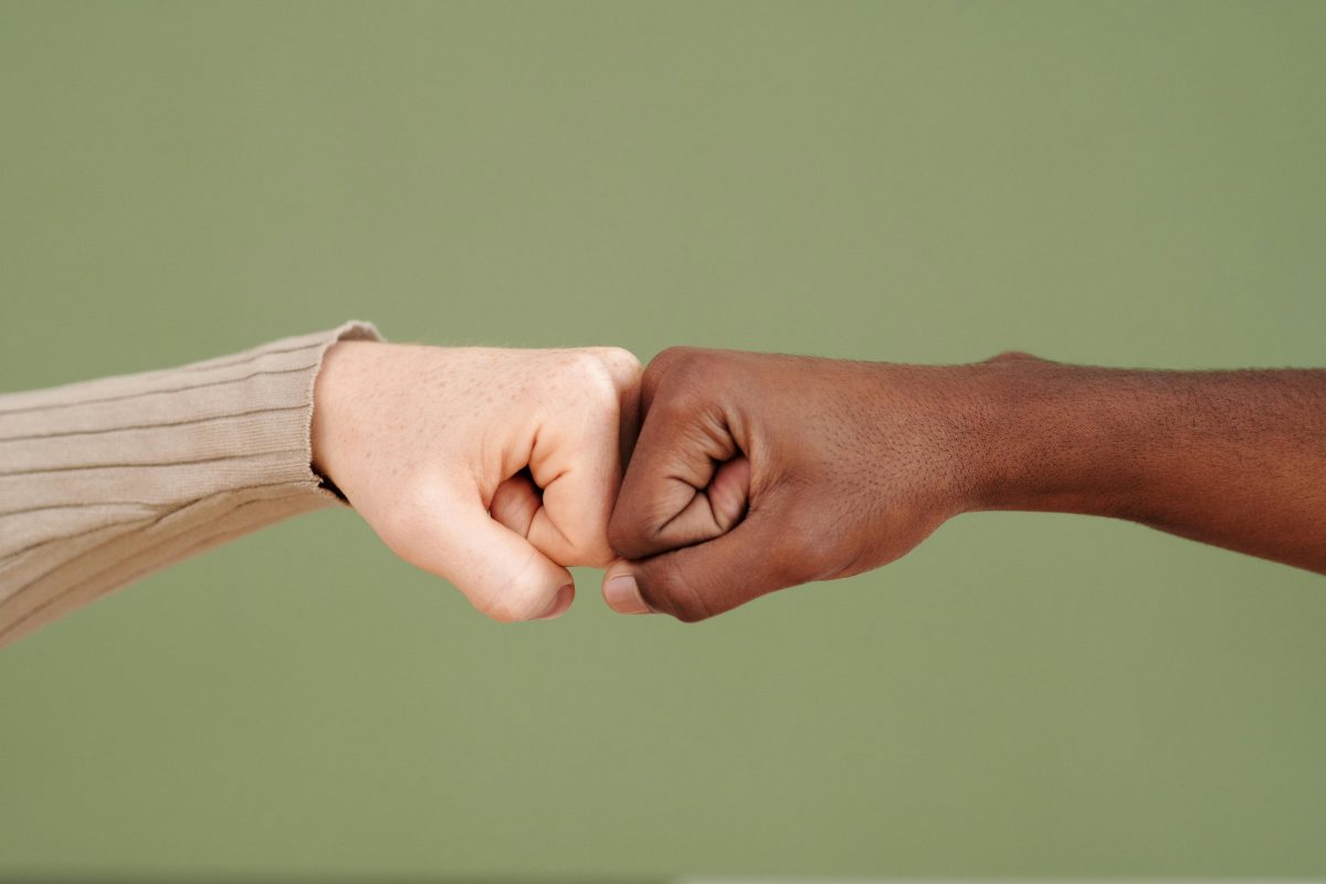 Two hands meet in the center of this image, in a fist bump. The hand on the left has a light skin tone and the hand on the right has a brown skin tone.