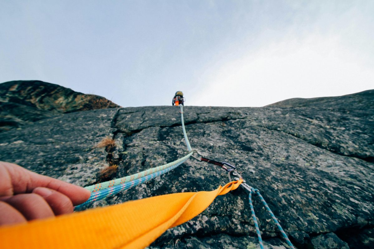 A hand holds a belay rope as a climber scale a cliff face.