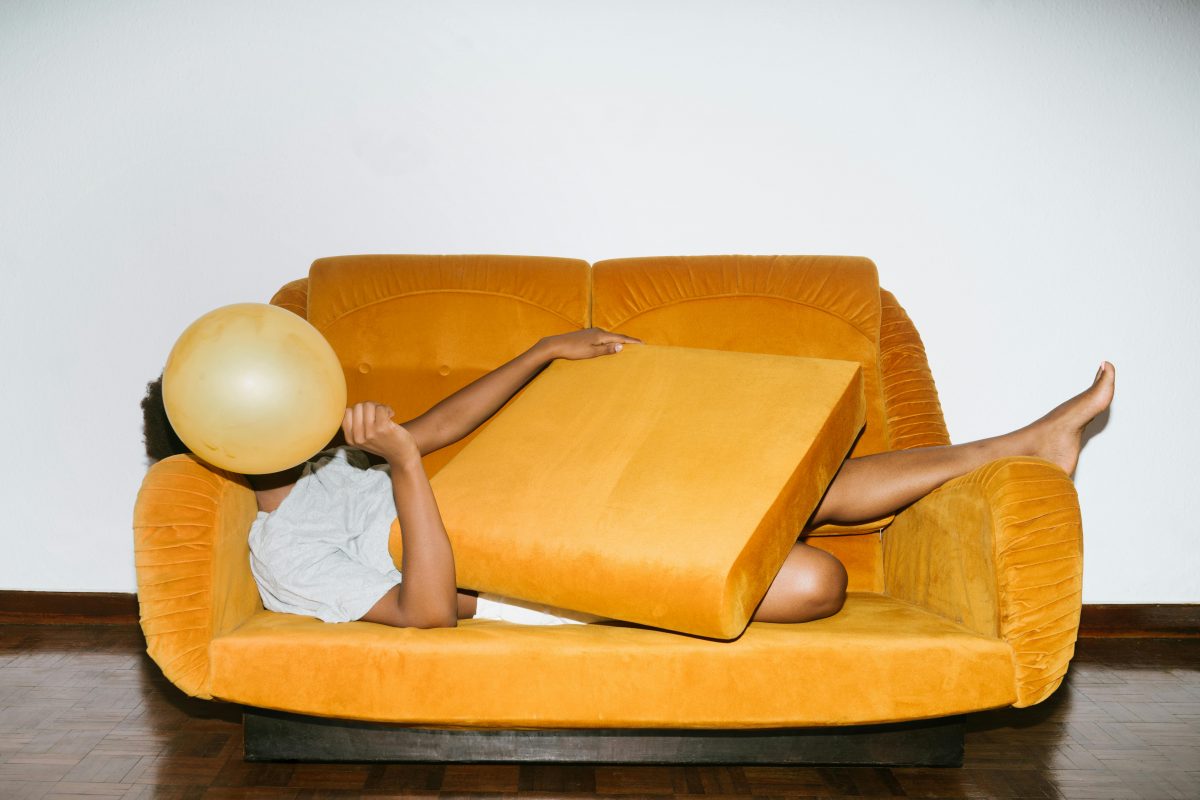 I person naps on an orange couch, with a cushion in their lap, and a yellow balloon obscuring their face.