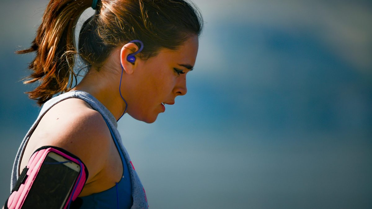 A young, light-skinned woman with wavy brown hair in a ponytail looks down after exercising. She is wearing earbuds and has a smartphone strapped to her arm.