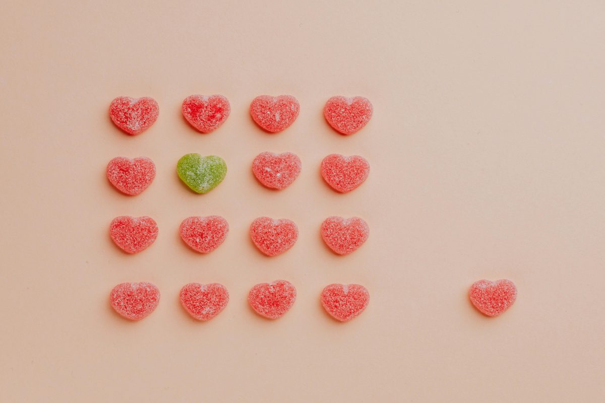 Amidst a group of red heart-shaped gummy candies, one is green. Another red one stands off to the side.