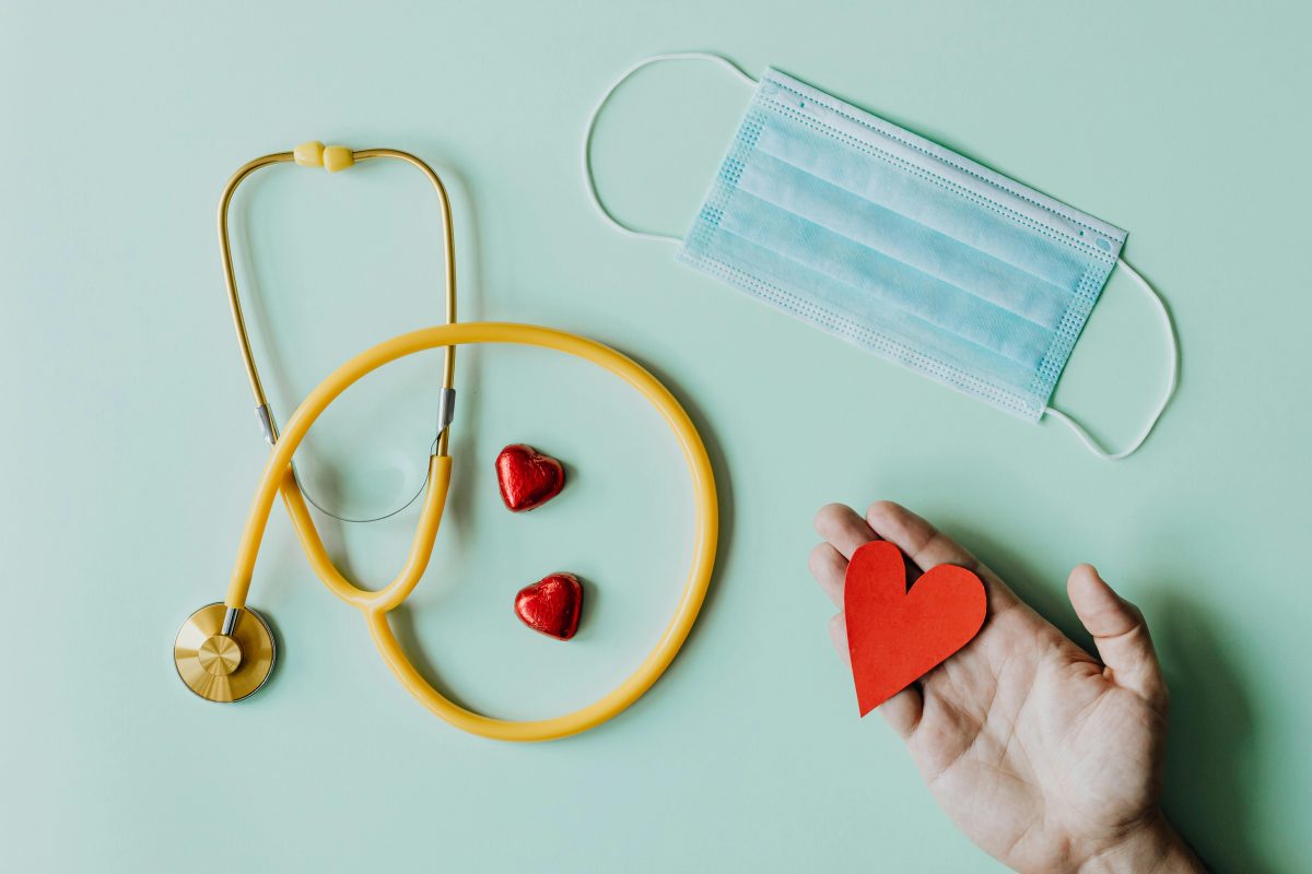 A yellow stethoscope is curled around two red heart shaped items. A blue surgical mask is to the right of it, and below the mask is a hand with a red paper or felt heart cutout.