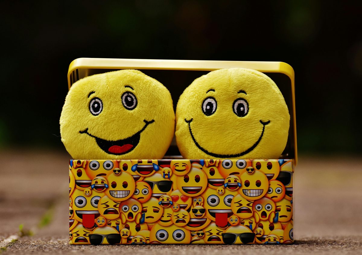 Two smiling emoji plushies sit inside of a lidded box covered with emojis.