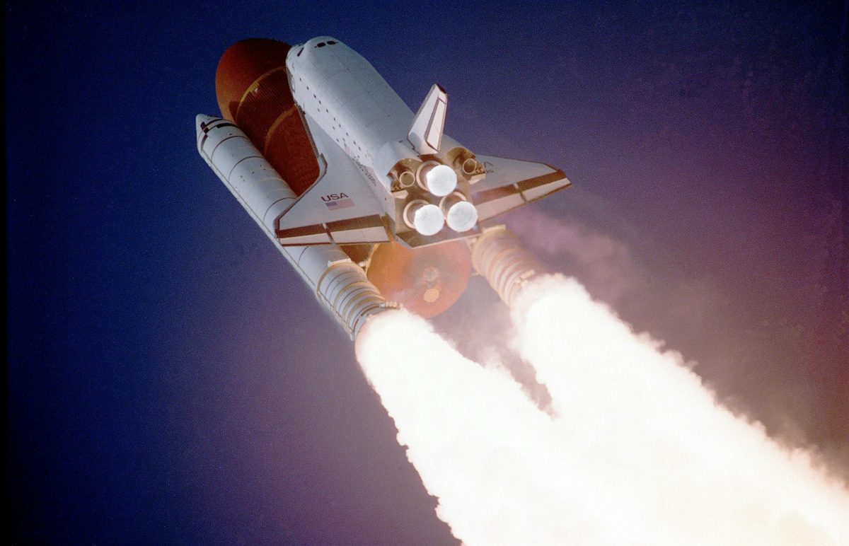 A space shuttle takes off against a dark blue sky. The flames shoot out from the rockets.