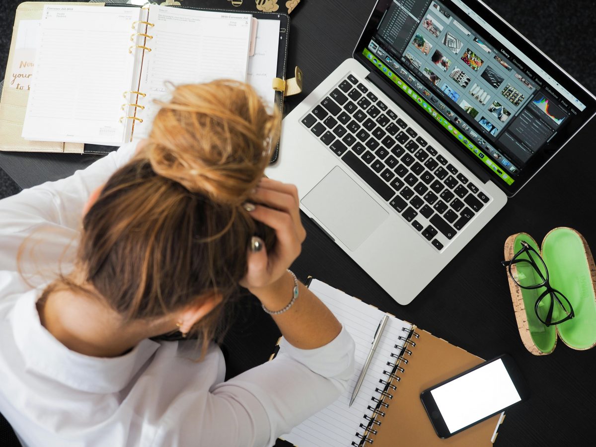 A young woman with light skin and brown/blond hair rests her head in her hand above her work desk, which holds a laptop, a planner, a notebook and a phone.