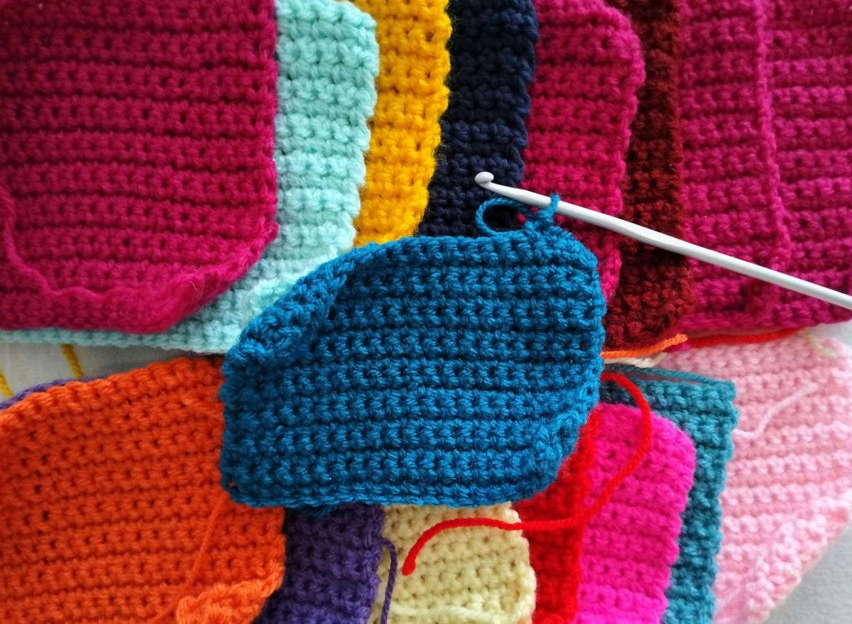 This photo shows layers of crocheted squares in many colors. In the center, a crochet hook is in a loop of yarn on a partially finished blue square.