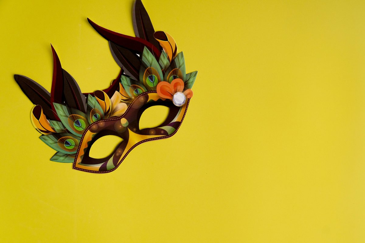 A carnival mask covered in colorful feathers lies on a yellow background.