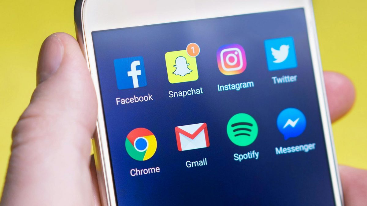 A photo of a hand holding a white smart phone with social media app icons across the top of the screen. The apps include Facebook, Snapchat, Instagram, Twitter.