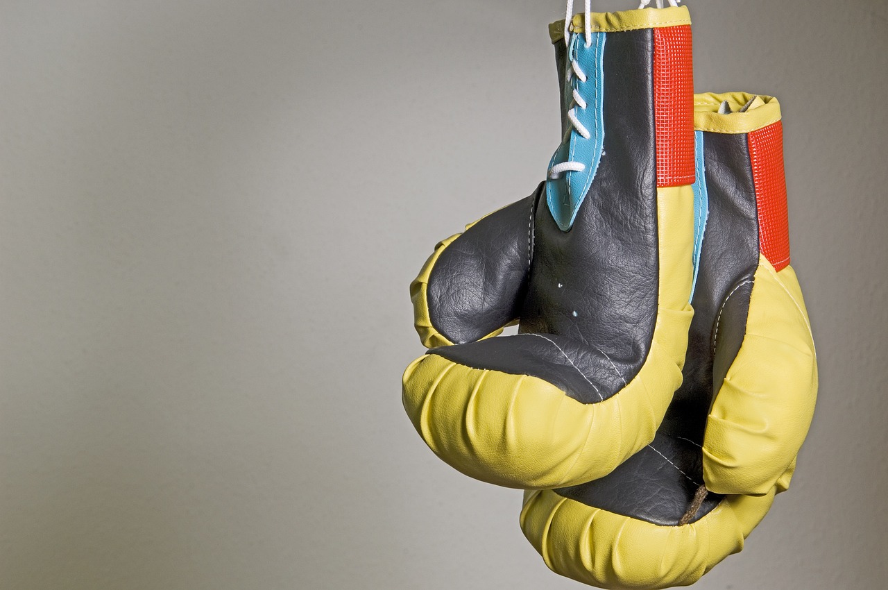 Two boxing gloves hanging down with a grey background. Gloves are black, yellow, red, and blue