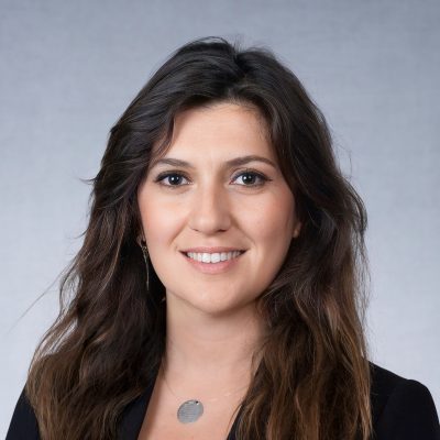 Portrait of Zeynep Akdemir. She is a woman with long, wavy dark brown hair and light skin. She is wearing a black suit jacket and a simple silver medallion necklace.